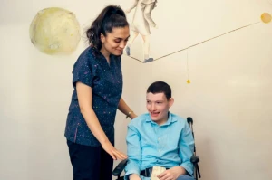 A caregiver stands beside an adult with a developmental disability using a wheelchair, offering friendly support and encouragement in a warm, home-like residential setting.
