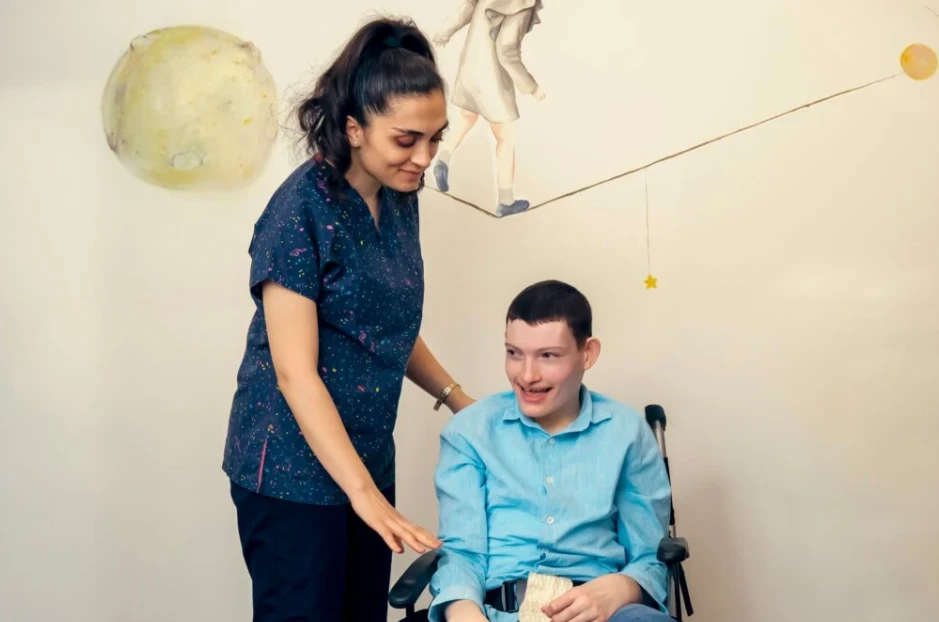 A caregiver stands beside an adult with a developmental disability using a wheelchair, offering friendly support and encouragement in a warm, home-like residential setting.