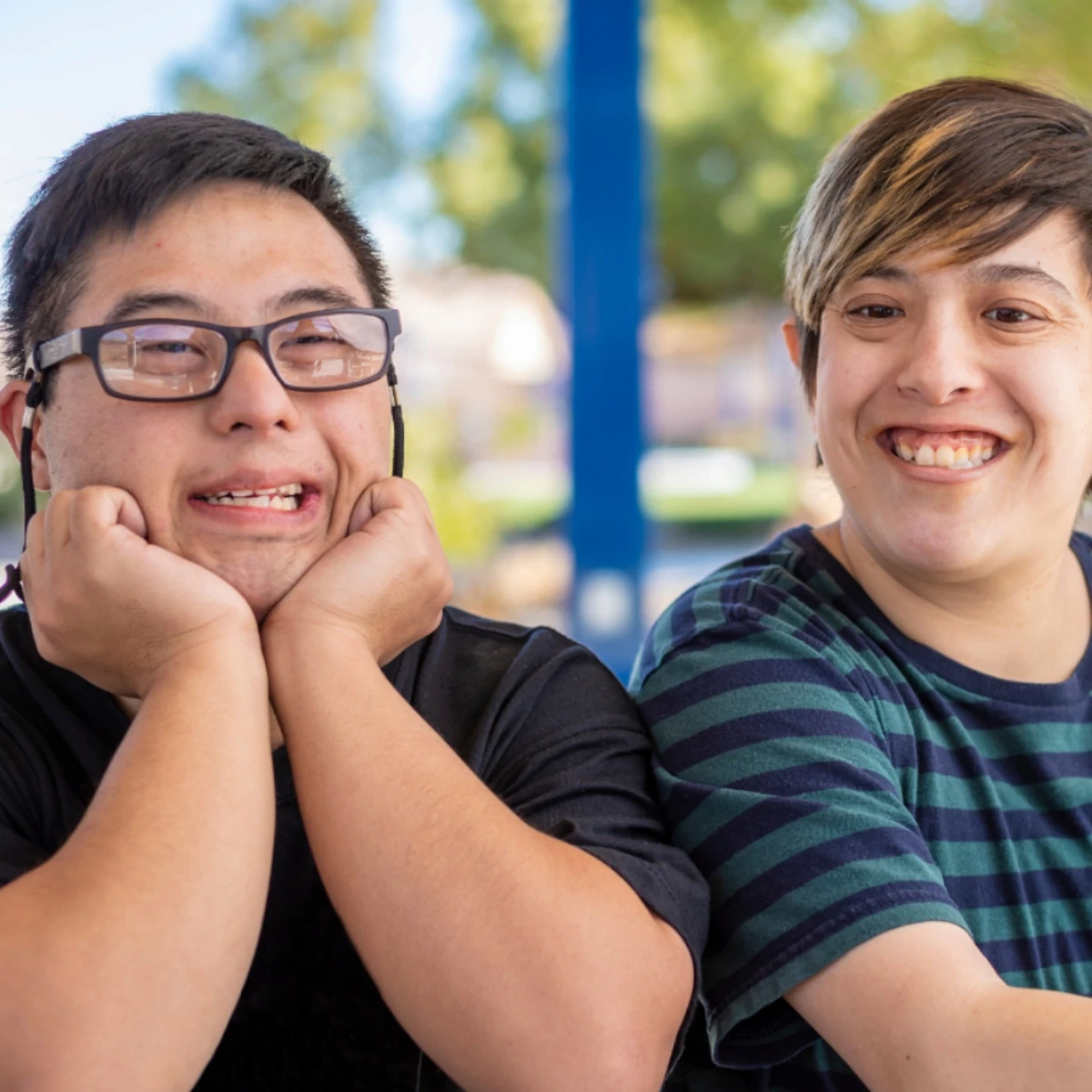 Two young men outdoors smiling warmly at the camera, one wearing glasses with his chin resting on his hands, both showing genuine joy and friendship in a community setting.